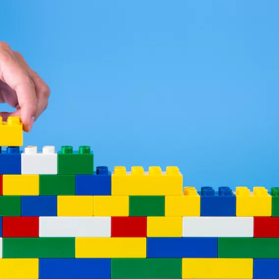 Photo of lego bricks of different colours stacked up with a hand adding another brick, against a blue background.
