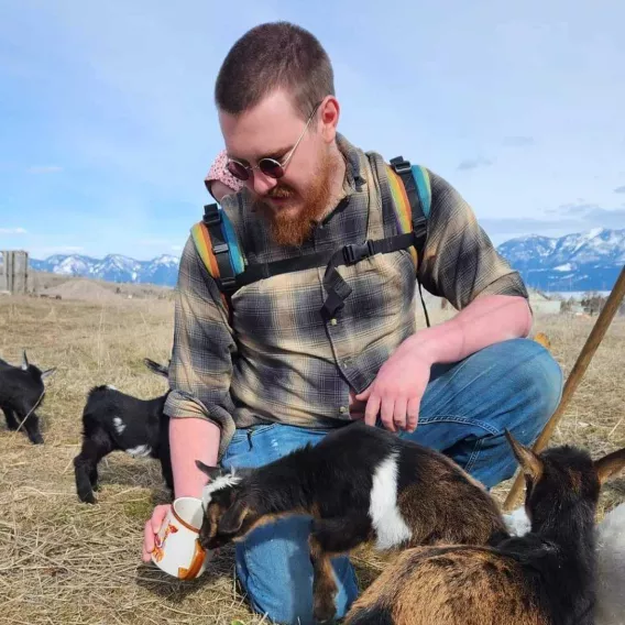 Photo of Isaiah, wearing a baby in a rainbow back carrier, holding a mug of tea which is being sniffed at by a baby goat.