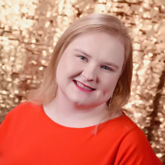 Photo of Kristen Goode, a Caucasian woman with shoulder-length, straight blonde hair and blue eyes. Kristen is wearing an orange dress and silver dangling earrings. The background is gold sparkles.